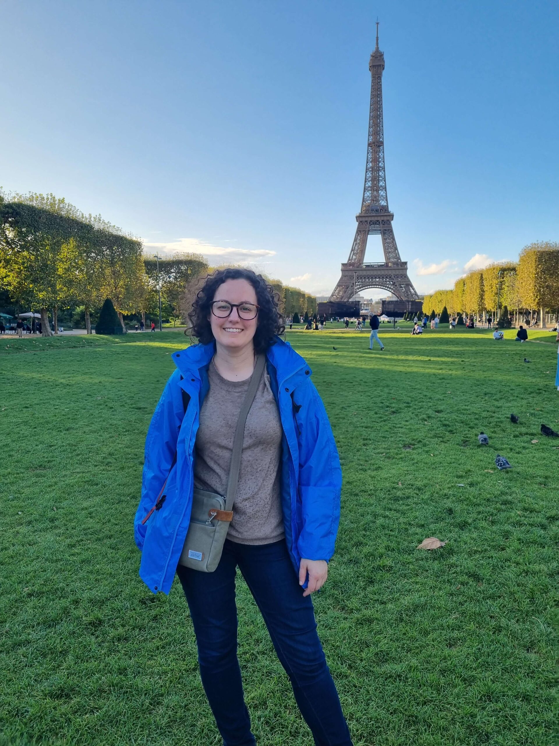 Charlotte is wearing a blue jacket, jeans and a brown top. She is standing on a grassy area in front of the Eiffel Tower, smiling.
