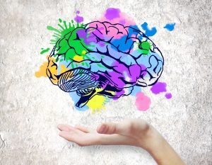 A human hand is gently open beneath a stylised, colourful illustration of a brain on a textured light background, representing creativity, diversity, and support for neurodivergent thinking.